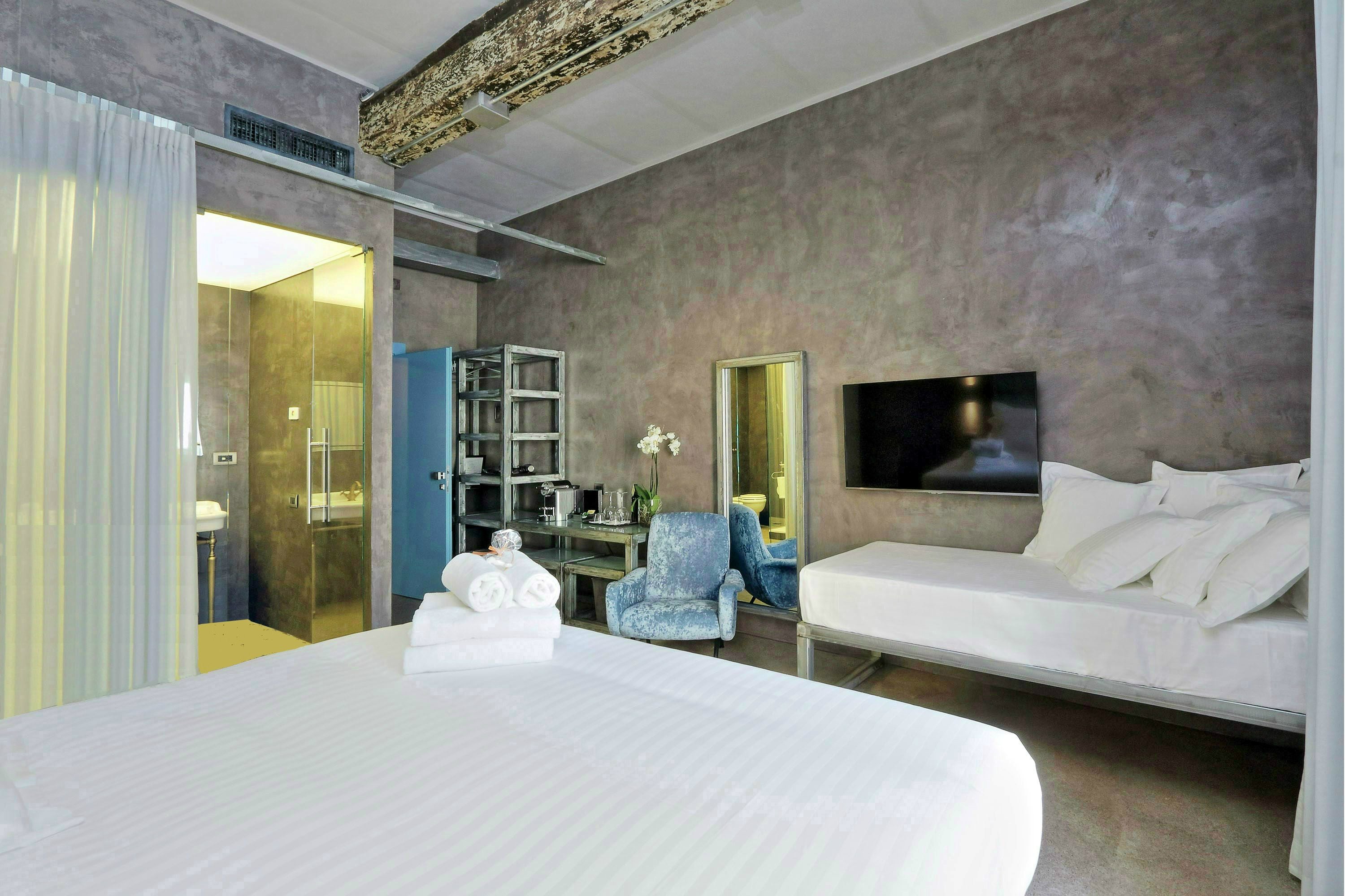 A modern hotel room with a large bed, a TV, a seating area with a chair, and a bookshelf.