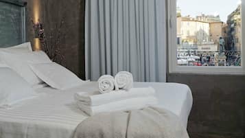 A neatly made bed with white linens and towels, a view of a cityscape through a window, and a decorative branch on the bedside table.