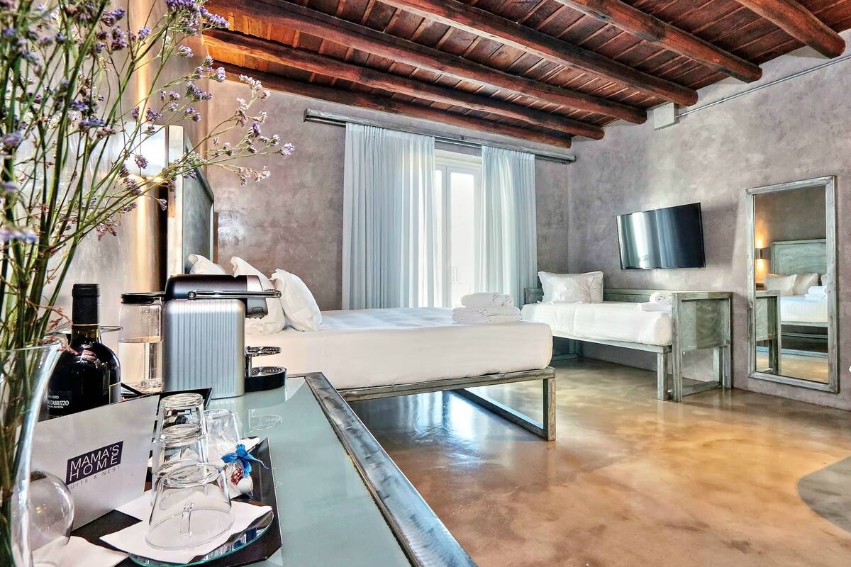A modern hotel room with a bed, a sofa, a TV, and a dining area with a tray of glasses and a bottle.