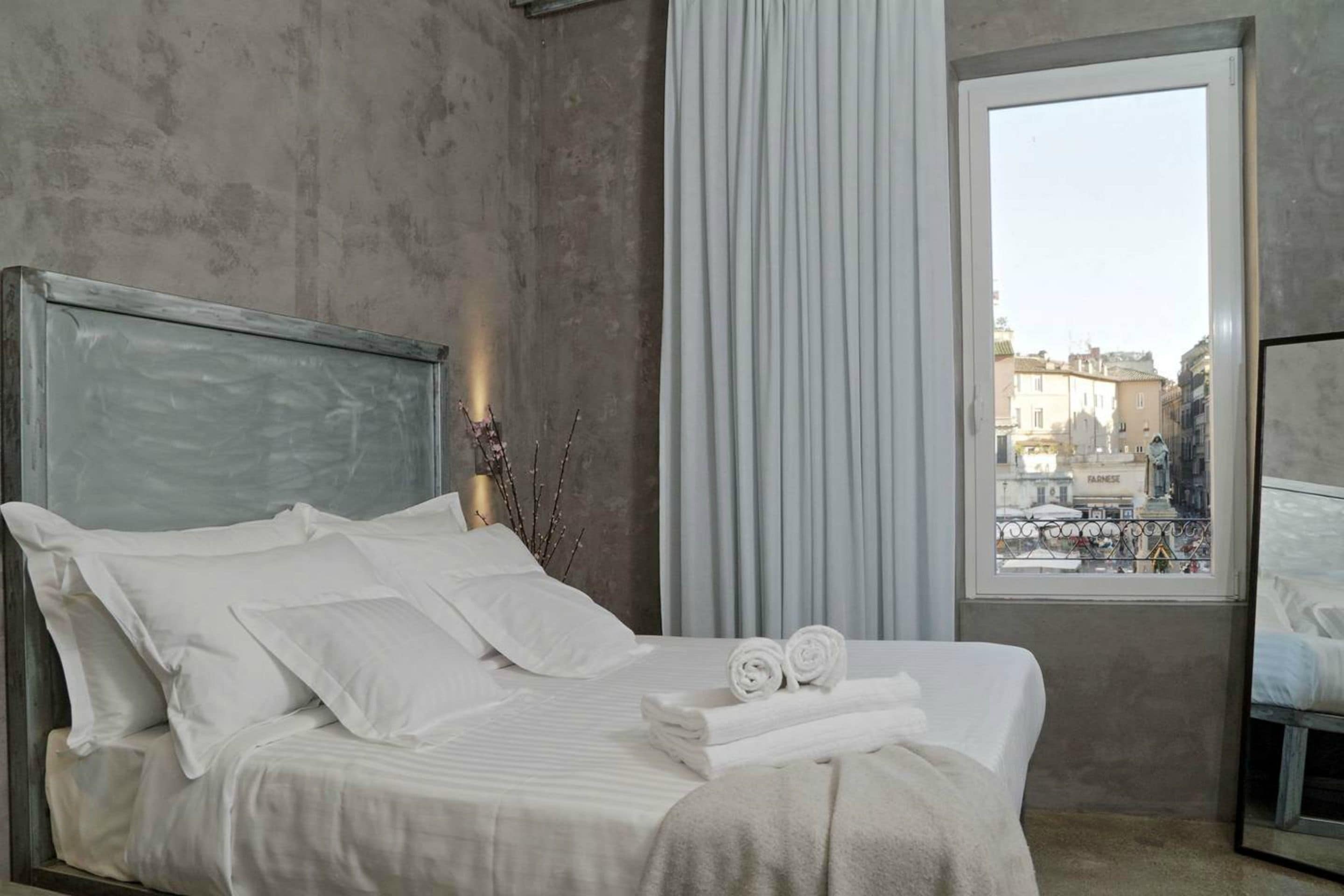 A neatly made bed with white linens and a headboard, a window with a view of buildings, and a curtain to the side.