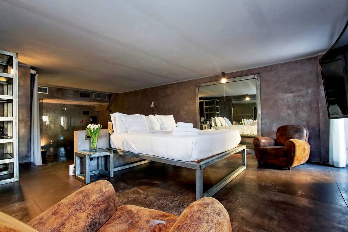 A modern bedroom with a large bed, a side table with a vase of flowers, and a leather armchair.