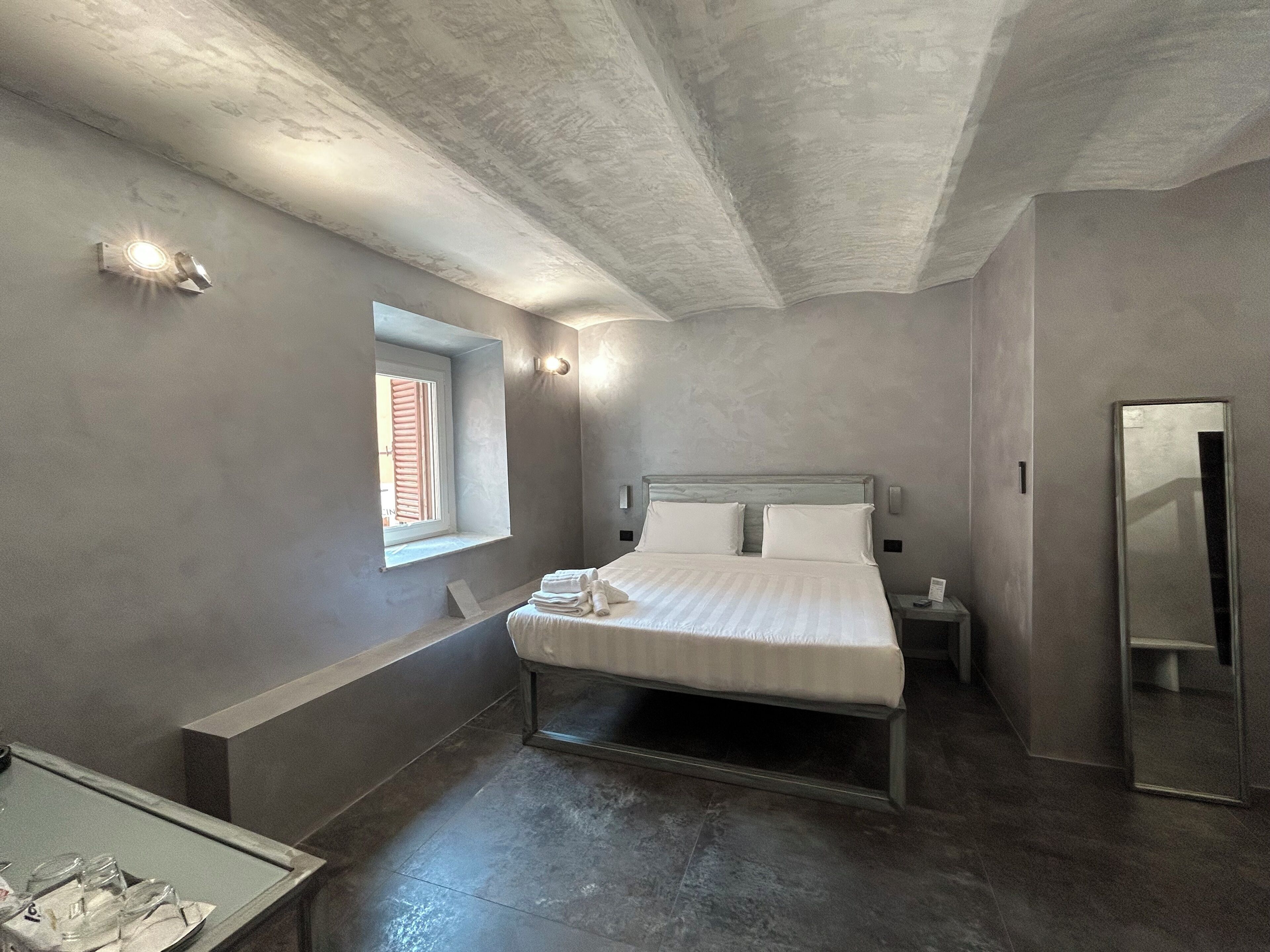 A modern bedroom with a large bed, a small window, a bedside table, and a mirror.