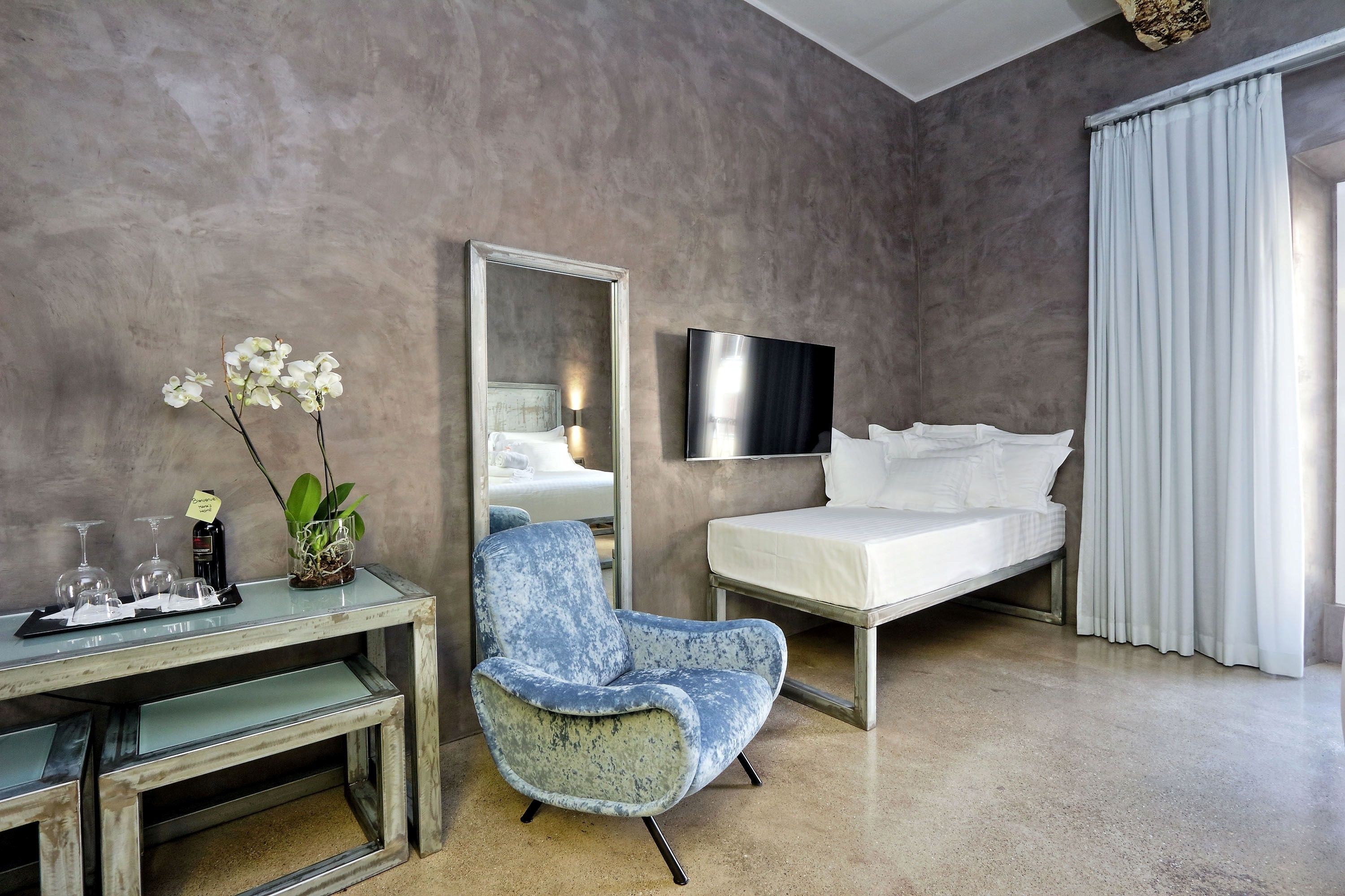 A modern hotel room with a bed, a desk with a chair, a mirror, and a potted orchid.