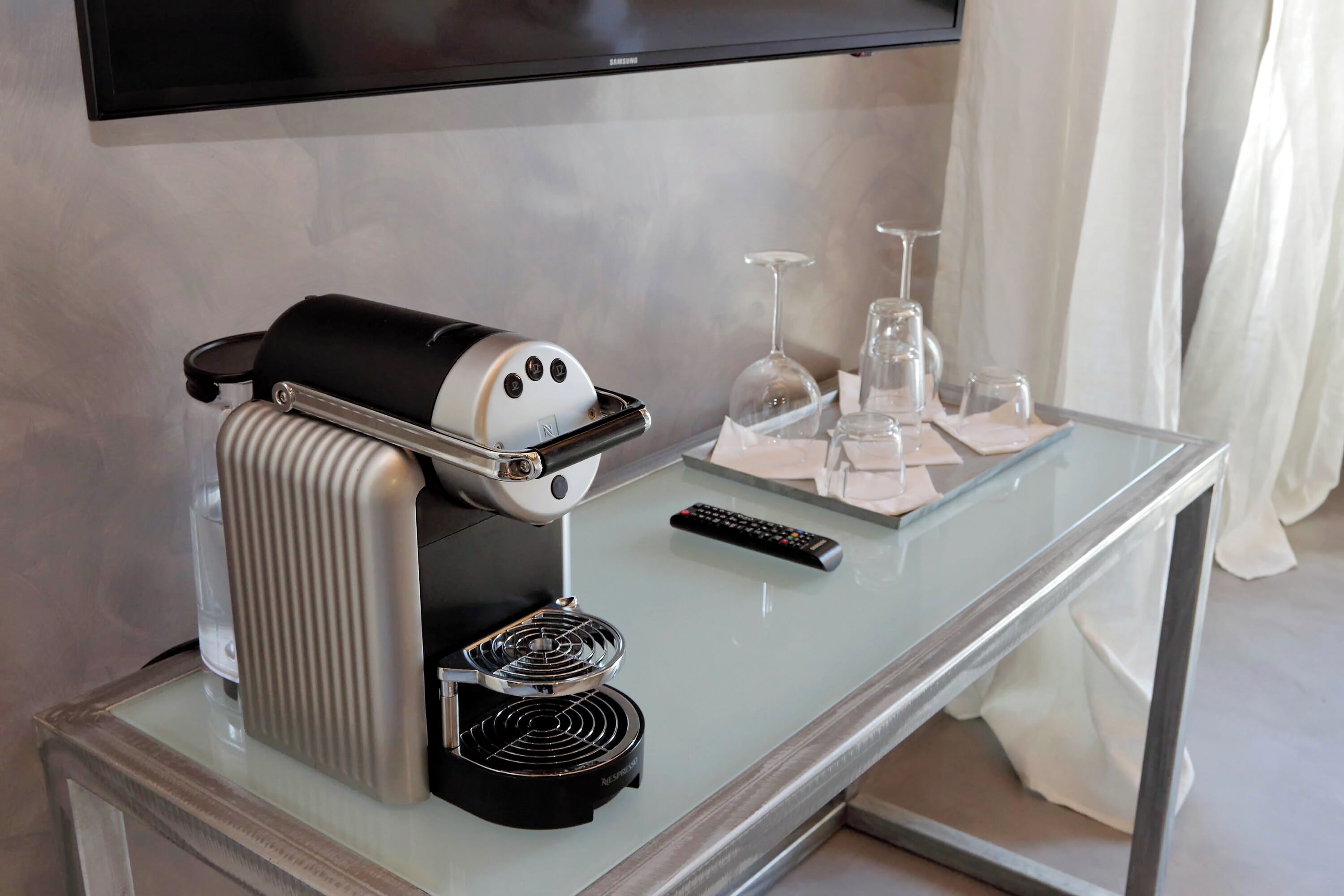 A modern hotel room with a glass coffee table, a coffee machine, a remote control, and a wine glass set.