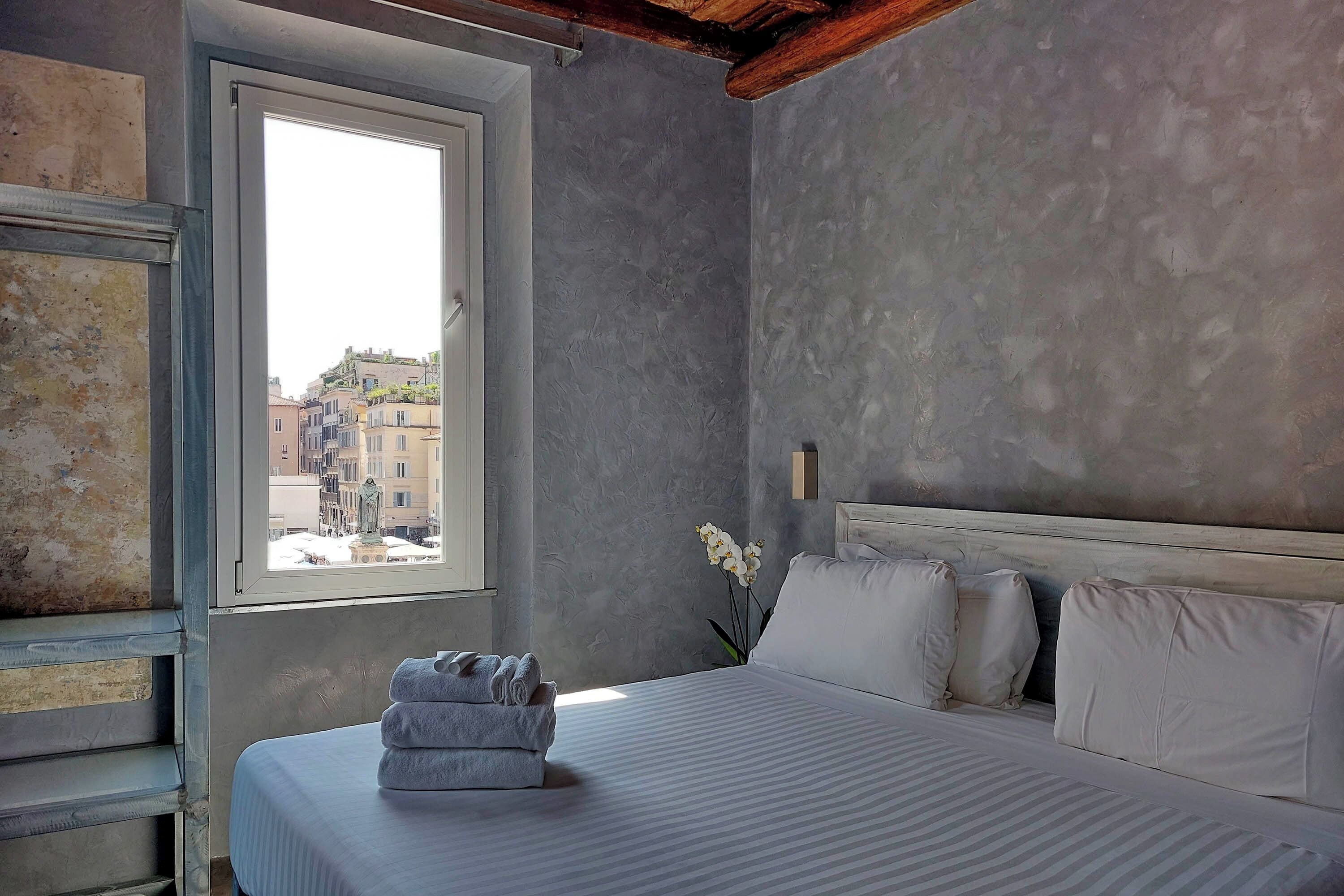 A bedroom with a bed, pillows, a window, and a view of buildings outside.