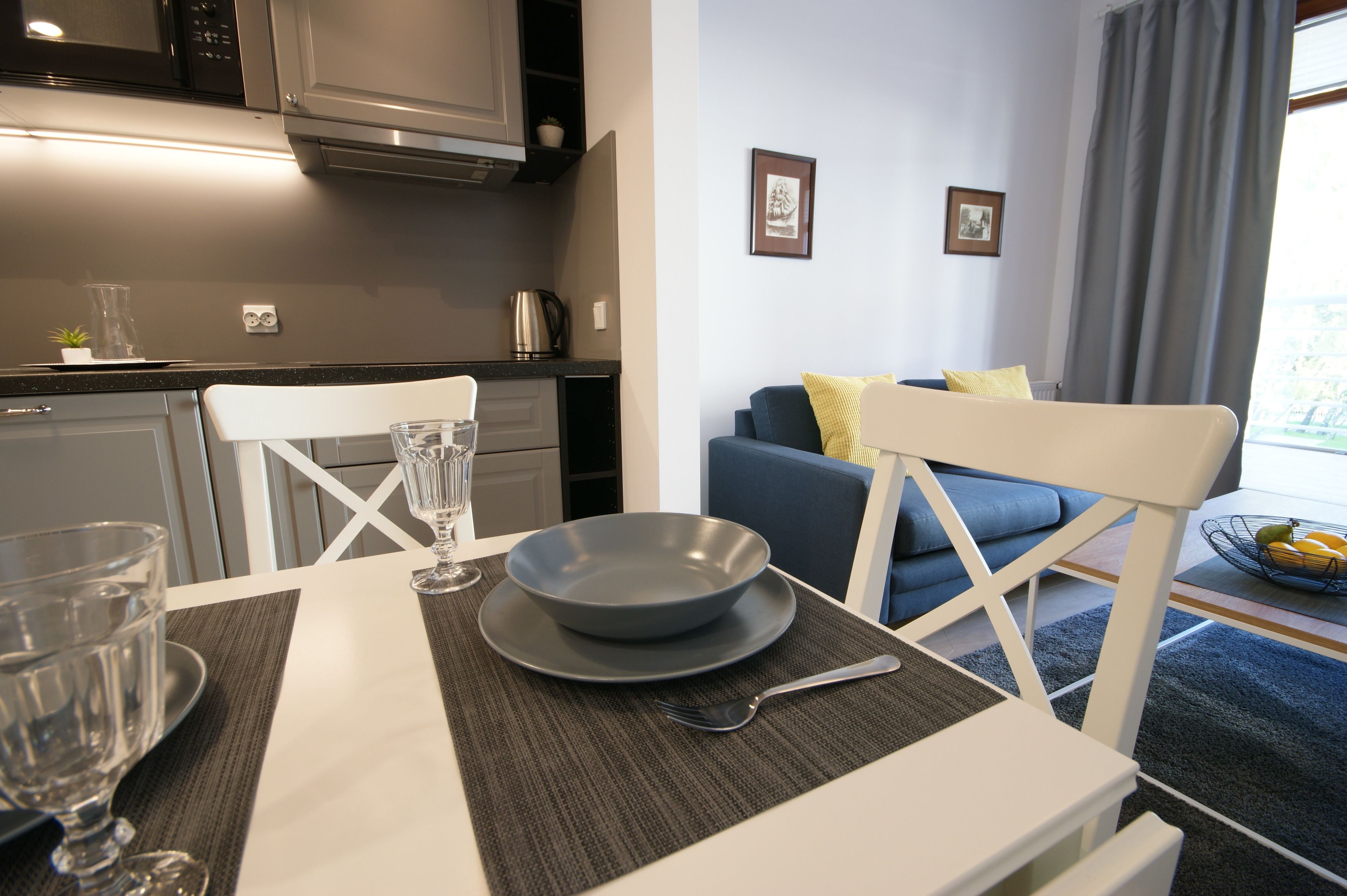 Un comedor con una mesa preparada para dos personas, una cocina al fondo y una sala de estar con un sofá y una ventana.