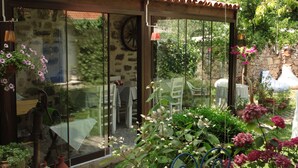 A glass-enclosed patio with a stone wall, outdoor furniture, and potted plants. - Istanbul Pansiyon (Ayvalik)