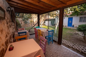 A covered patio area with a table and chairs, a stone wall, and a view of the garden. - Istanbul Pansiyon (Ayvalik)