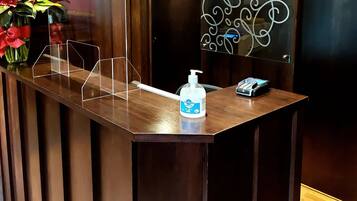 Un mostrador de recepción con una barrera de plástico transparente, dispensador de desinfectante para manos, un terminal de pago y un letrero decorativo.