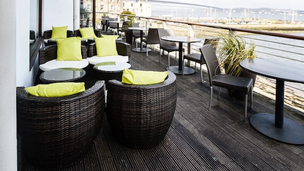 A terrace with wicker furniture, tables, and chairs, overlooking a waterfront with a bridge in the background.
