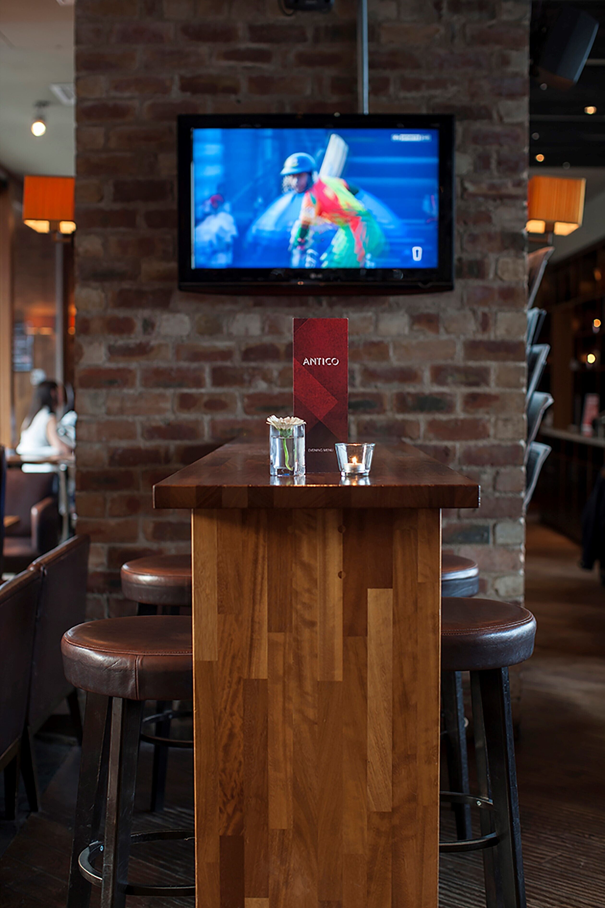 A bar area with a wooden counter, a TV mounted on a brick wall, and bar stools.