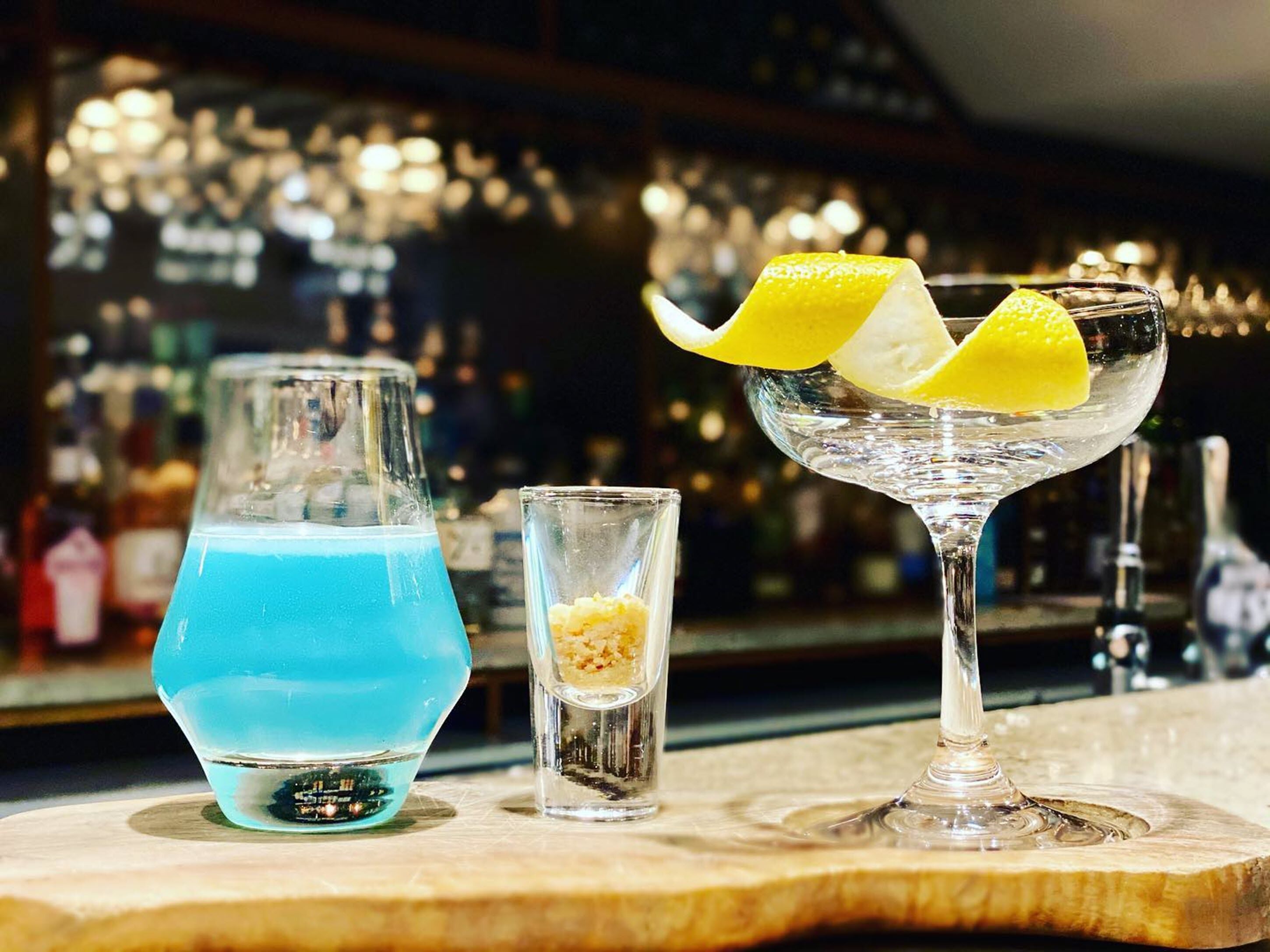 Três bebidas sobre um balcão de bar, com um fundo desfocado de garrafas e luzes.