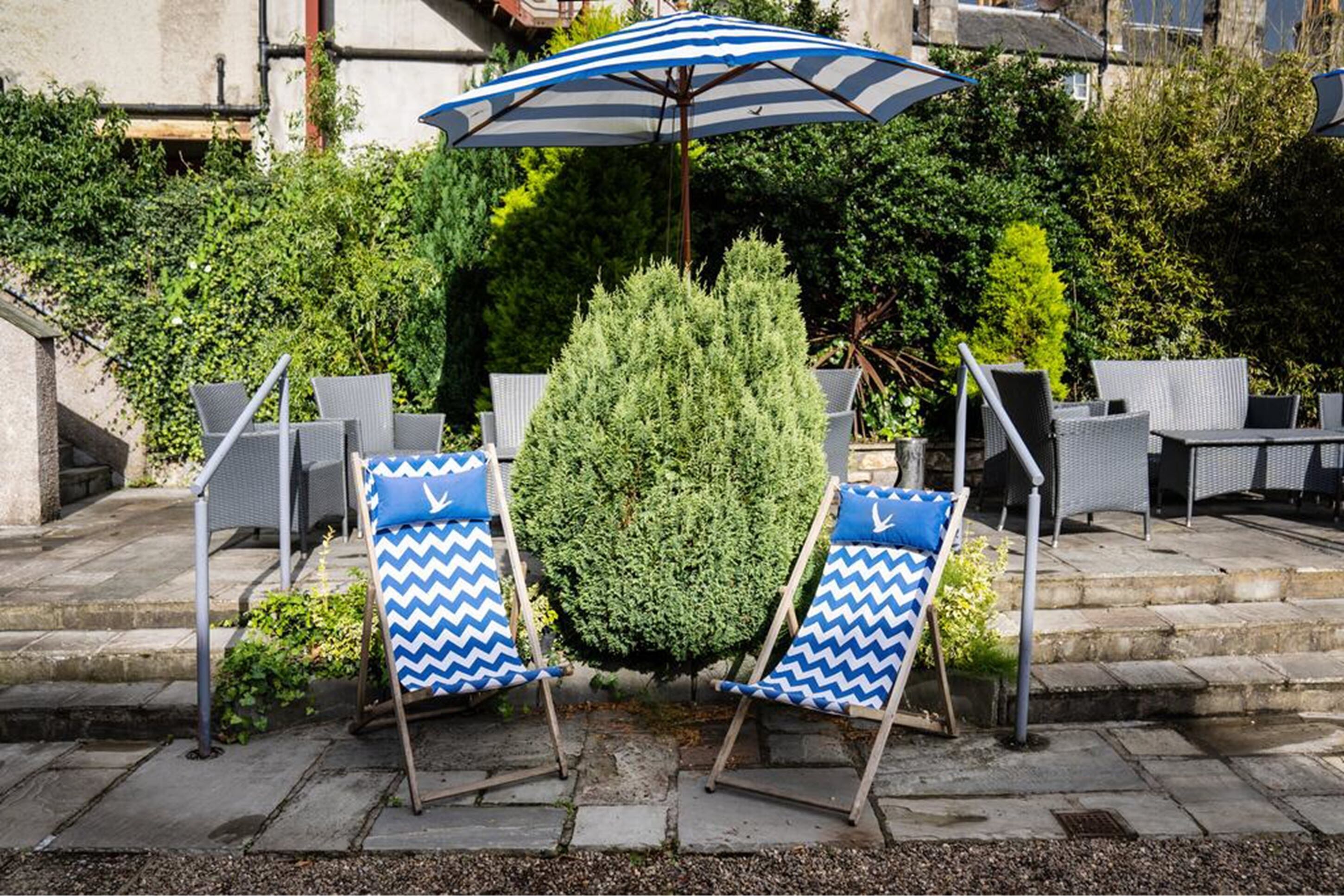 Deux fauteuils de jardin bleus et blancs à motifs chevrons, accompagnés d’un grand parasol rayé, d’un arbuste taillé avec soin et de meubles en osier, dans un espace patio extérieur.