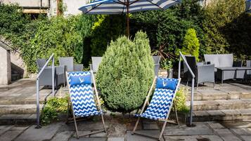Deux fauteuils de jardin bleus et blancs à motifs chevrons, accompagnés d’un grand parasol rayé, d’un arbuste taillé avec soin et de meubles en osier, dans un espace patio extérieur.