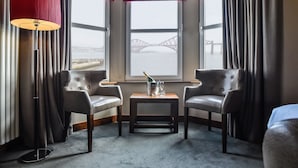 A hotel room with two leather armchairs, a small table with a bottle and glass, and a view of a bridge through large windows.