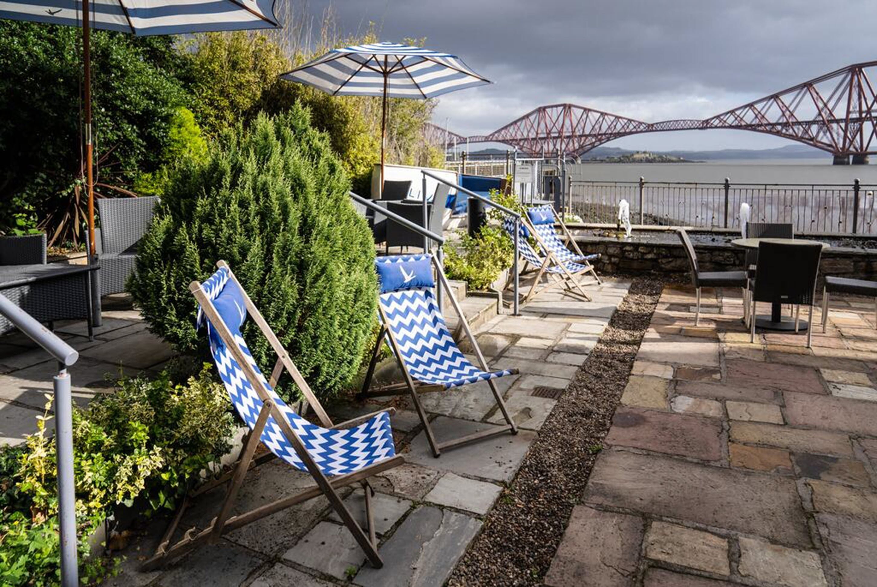 A patio area with blue and white striped umbrellas, lounge chairs, and a view of a bridge over water.