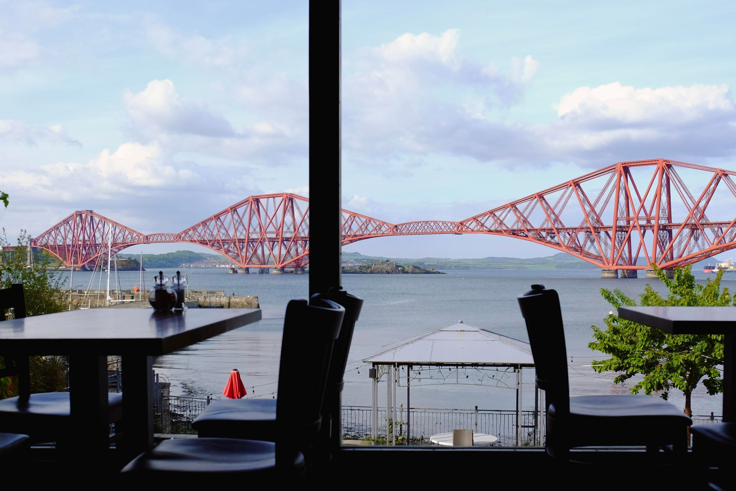Vista de un puente rojo sobre el agua, tomada desde un restaurante con mesas y sillas.