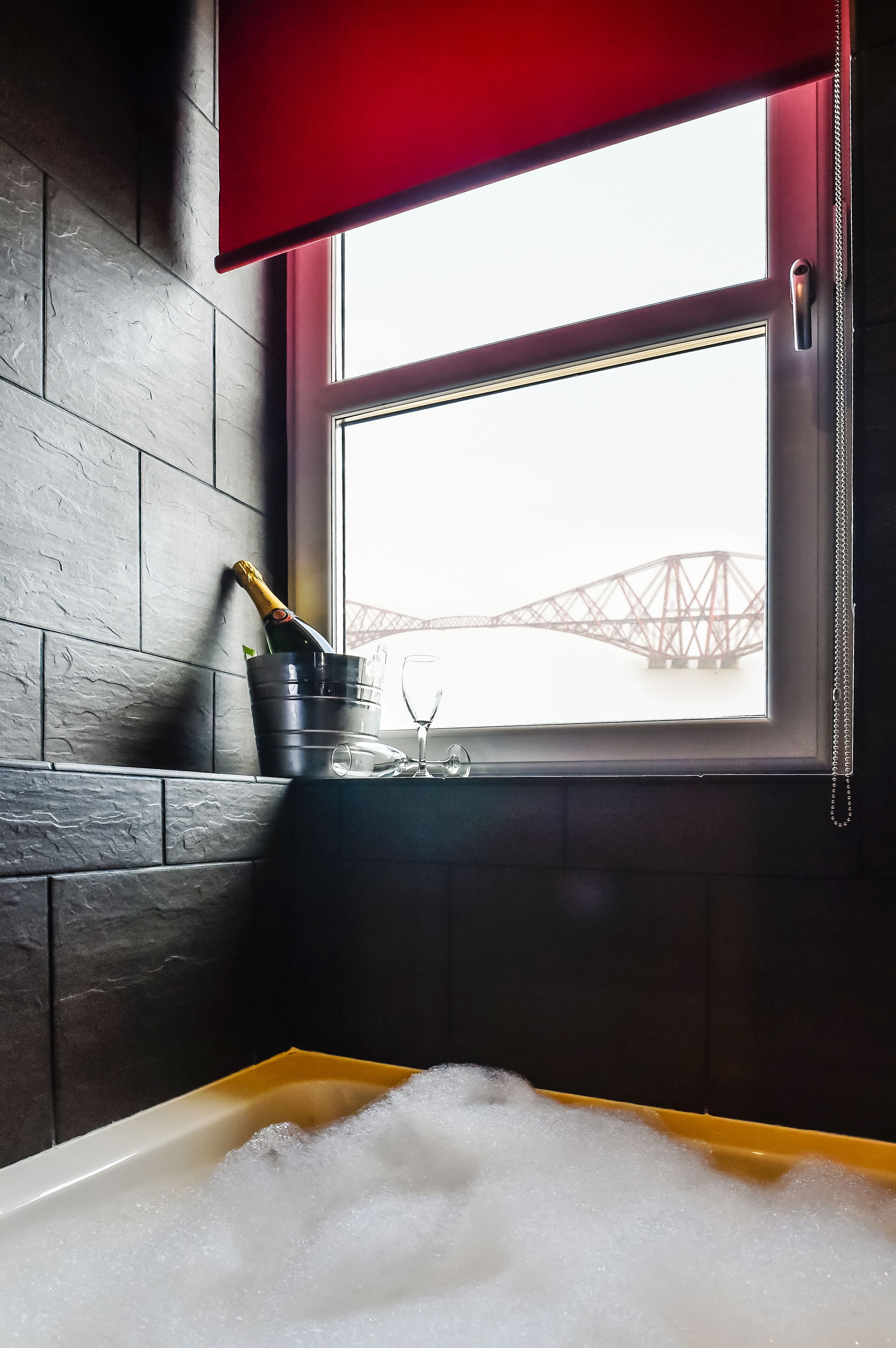 Una bañera llena de burbujas, un cubo con una botella y vasos, y una vista de un puente a través de una ventana.