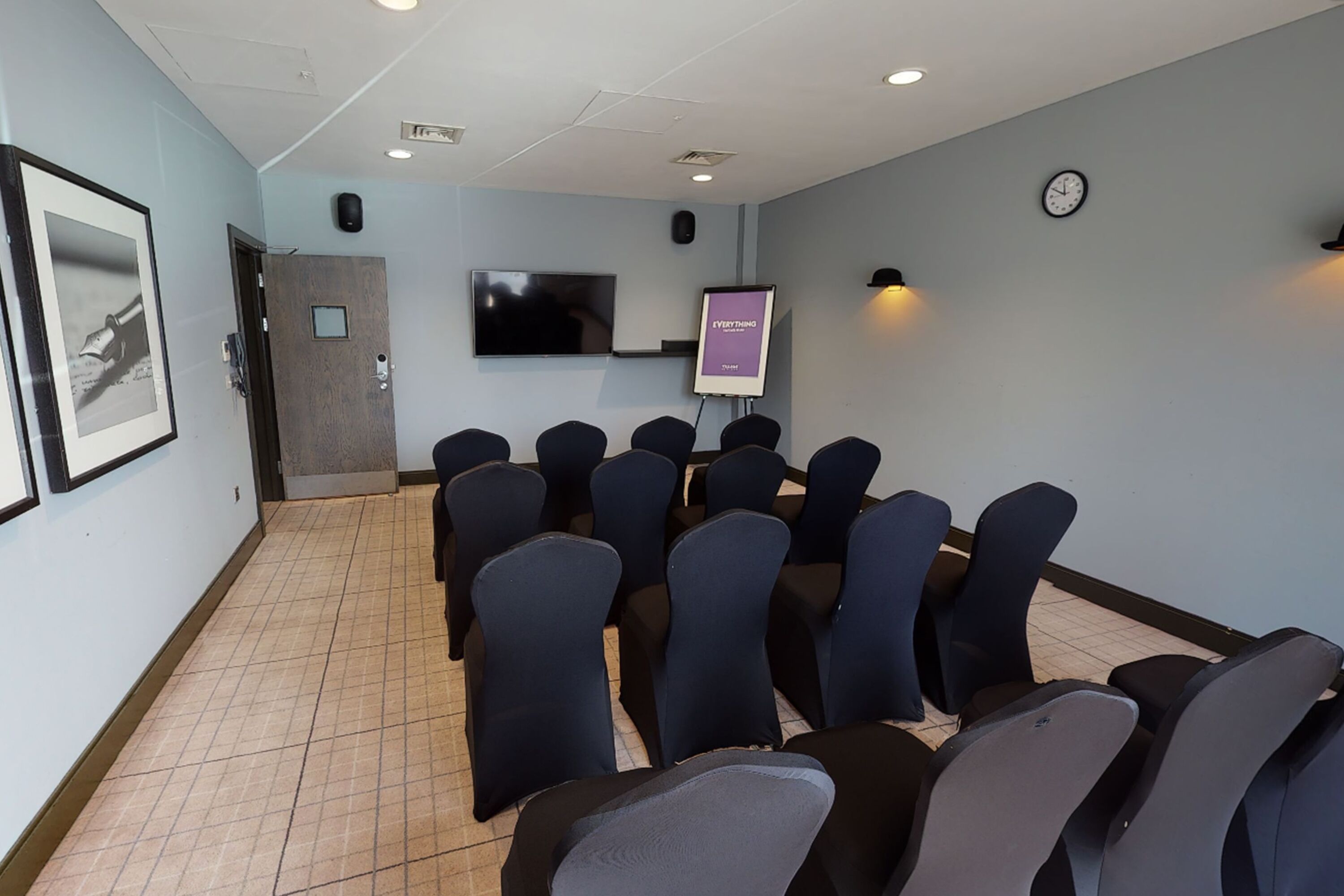 A conference room with rows of chairs, a projector screen, a clock, and a framed picture on the wall.