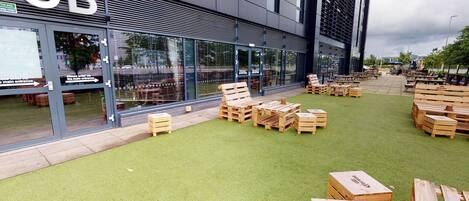 Un pub con terraza al aire libre sobre césped artificial, cajas de madera y fachada de edificio moderna.