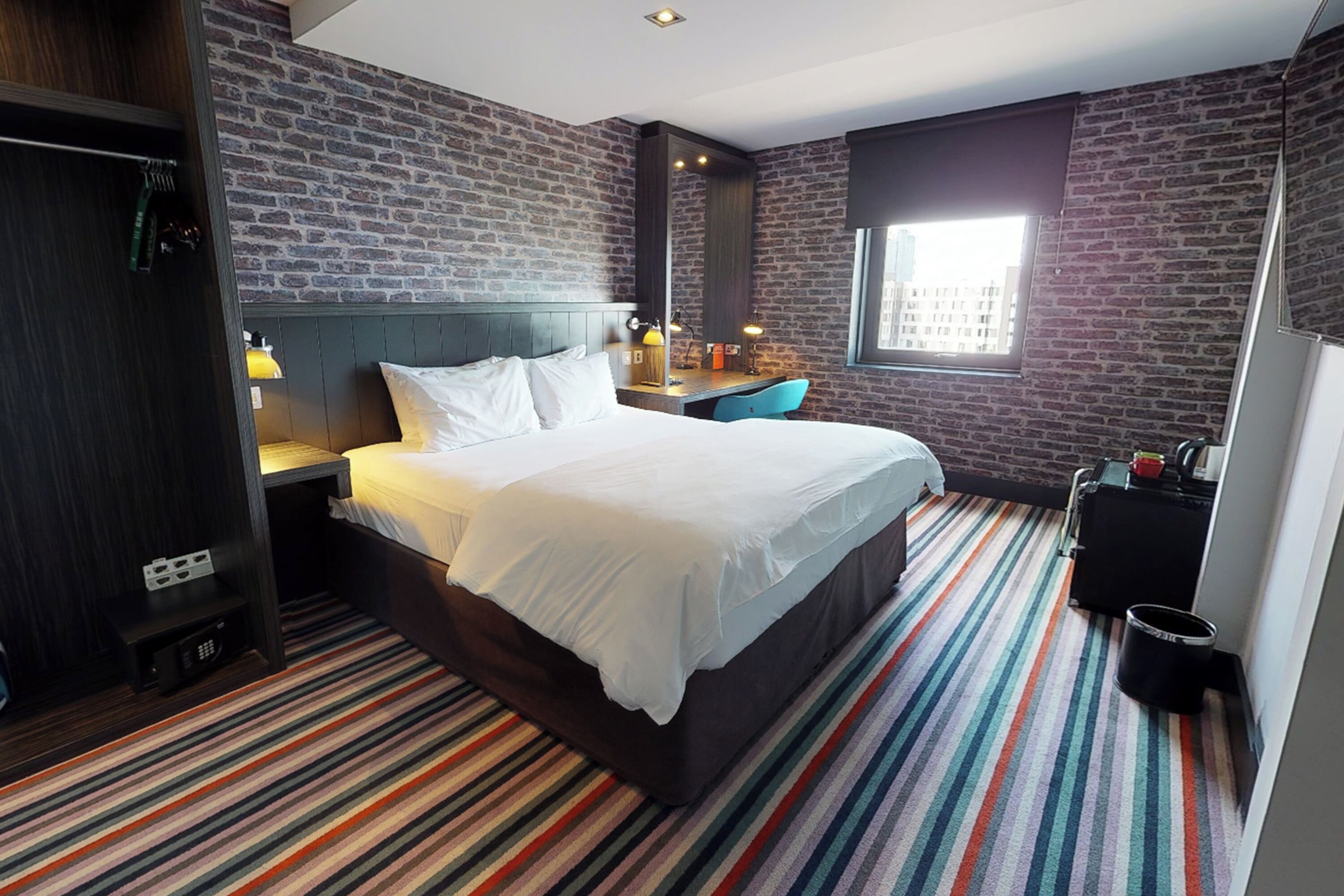 Ein Hotelzimmer mit Backsteinwand, einem Bett mit weißer Bettwäsche, einem Schreibtisch mit Stuhl und einem Fenster mit Blick auf Gebäude.