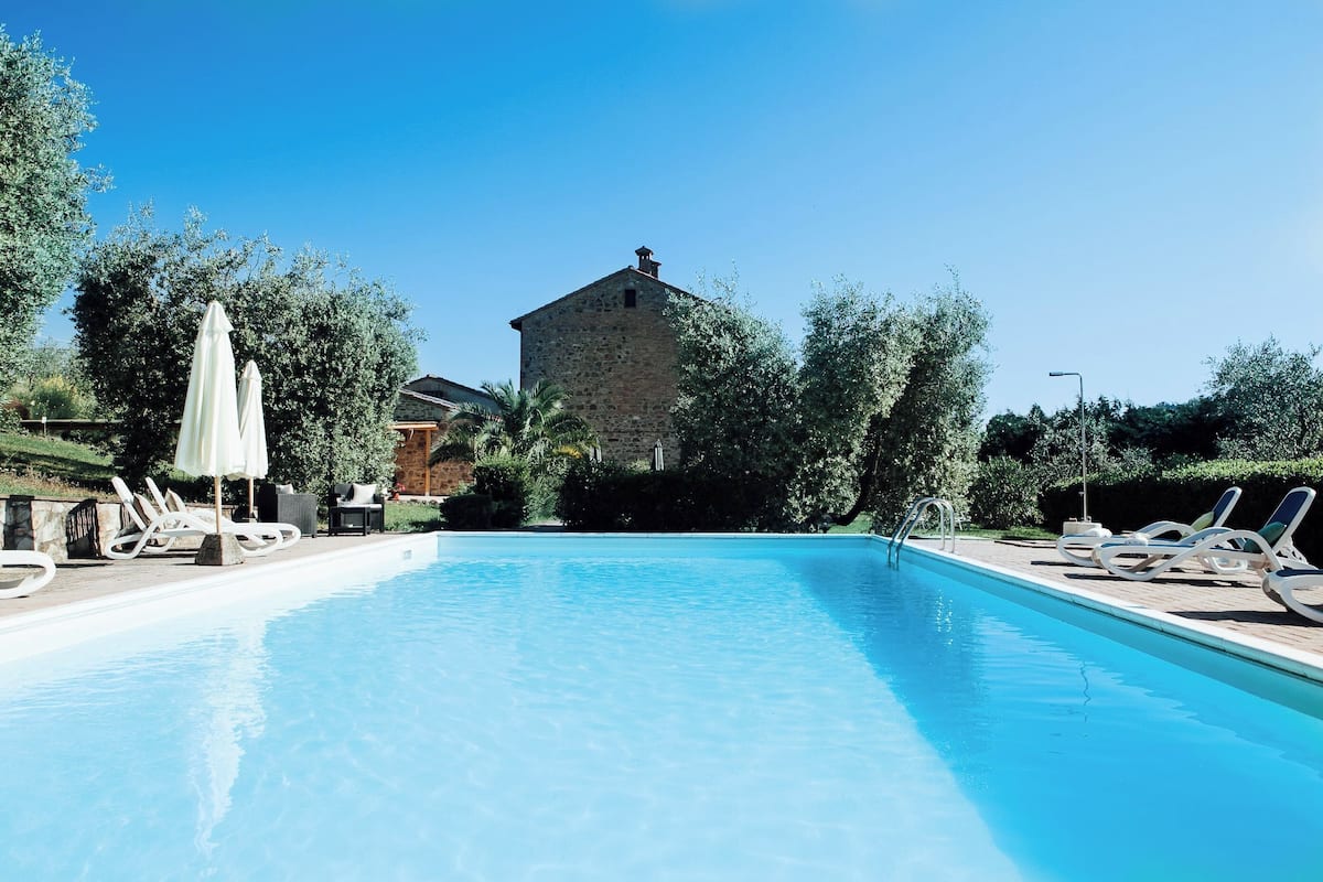 Ein Swimmingpool mit Liegestühlen und Sonnenschirmen, umgeben von viel Grün und einem Steinhaus im Hintergrund.