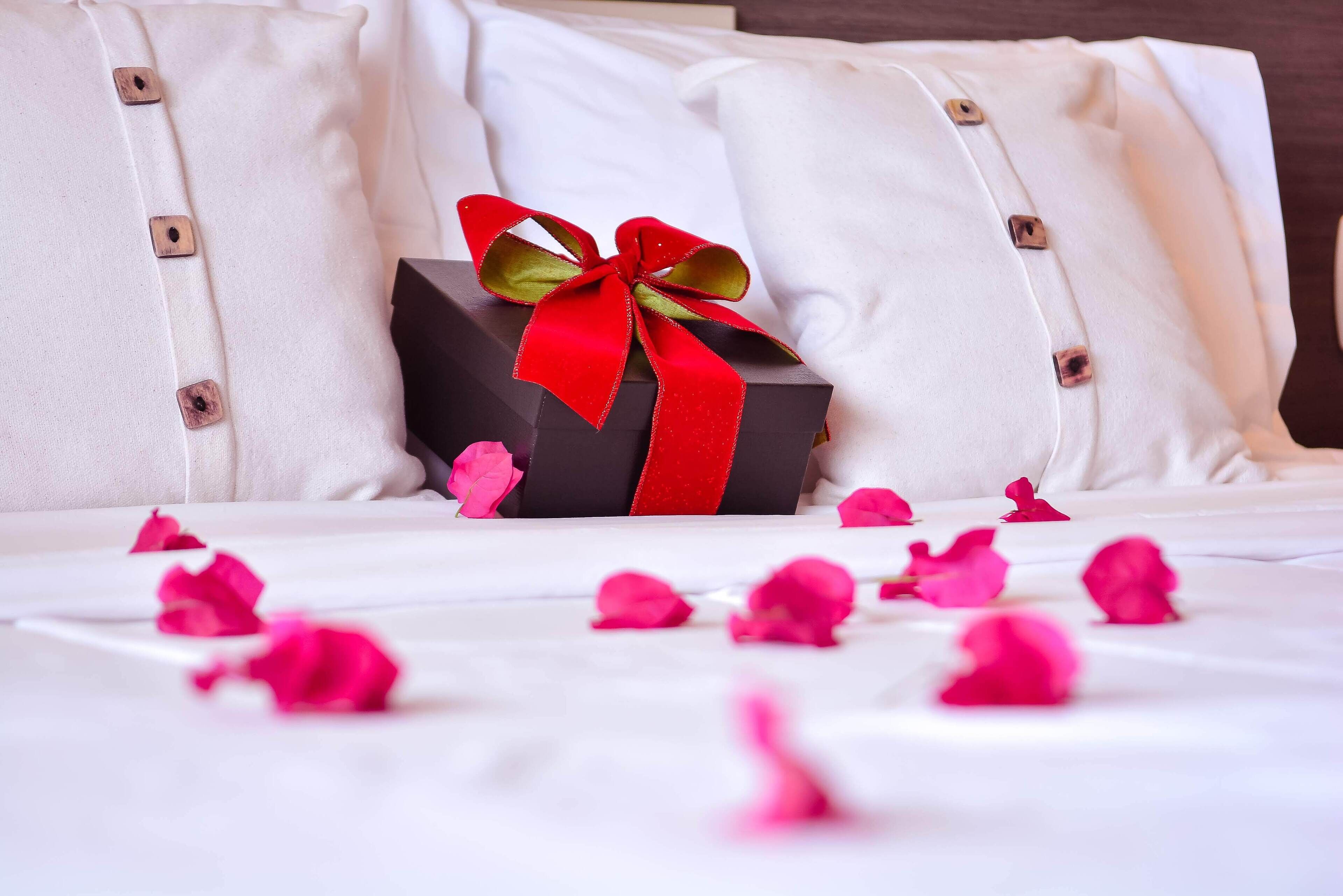 Una cama con ropa de cama blanca, una caja de regalo negra con un lazo rojo y pétalos de rosa rosados esparcidos.
