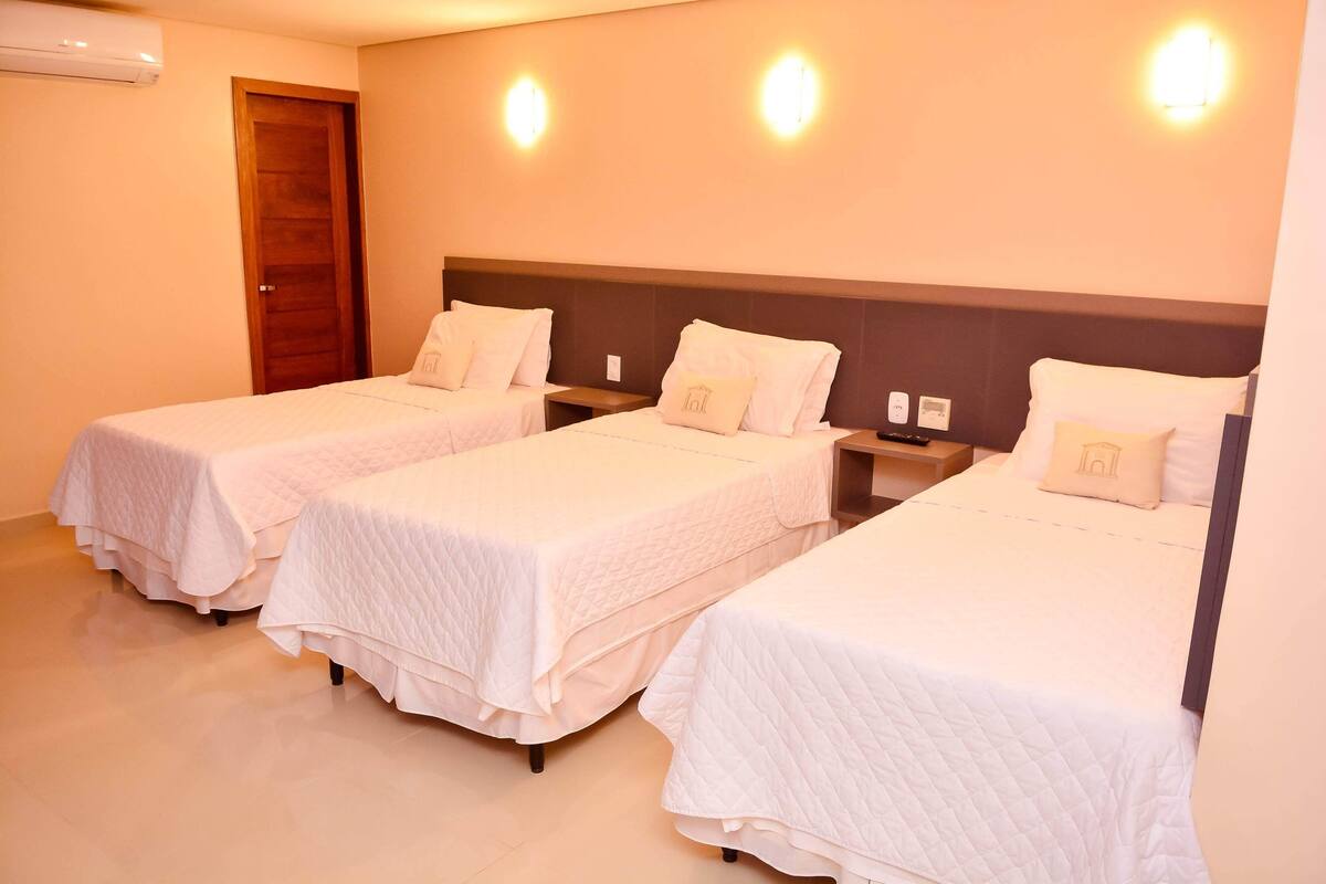 Une chambre d’hôtel avec trois lits simples, chacun recouvert d’une courtepointe blanche et agrémenté d’oreillers assortis. La chambre est dotée d’une porte en bois et d’un climatiseur fixé au mur.