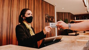 Una recepción de hotel con un panel de pared de madera, una recepcionista sosteniendo una tarjeta de presentación y un ordenador portátil sobre el mostrador.