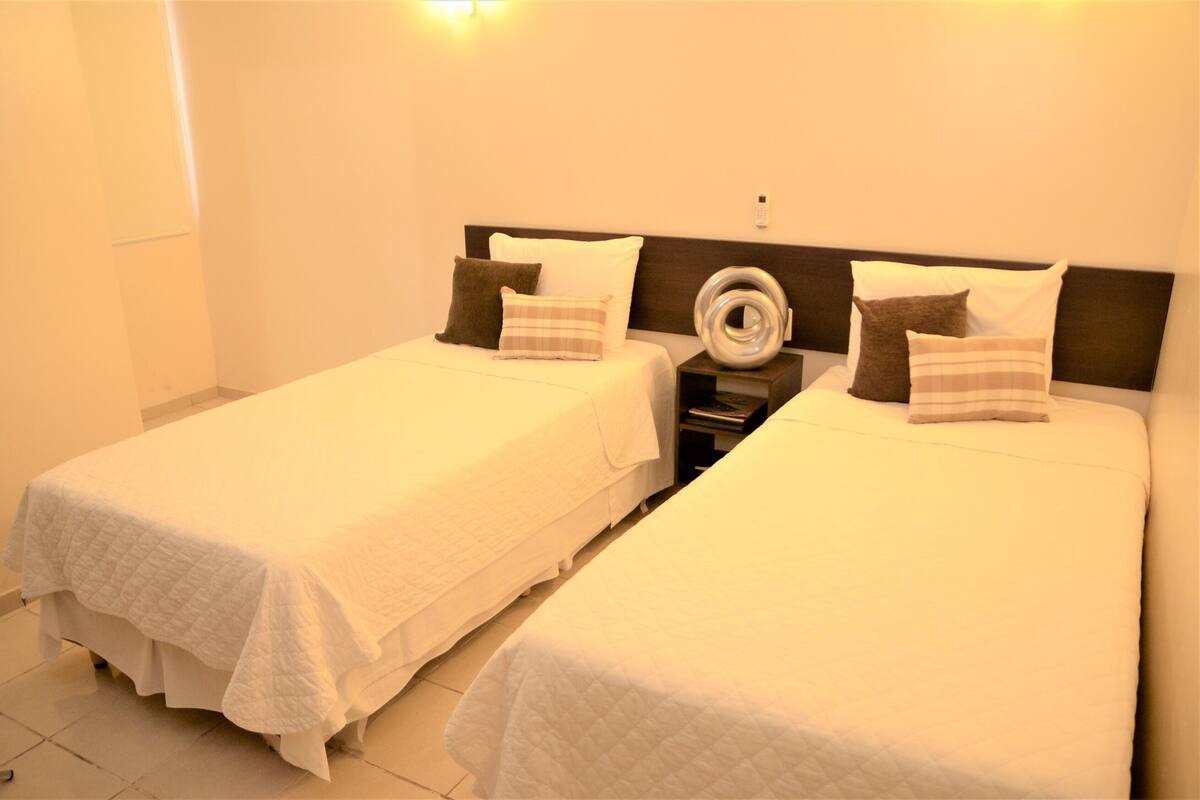 Une chambre d’hôtel avec deux lits simples, chacun recouvert d’une courtepointe blanche et agrémenté de plusieurs oreillers. On peut voir un objet rond et blanc sur la table de chevet, entre les deux lits.