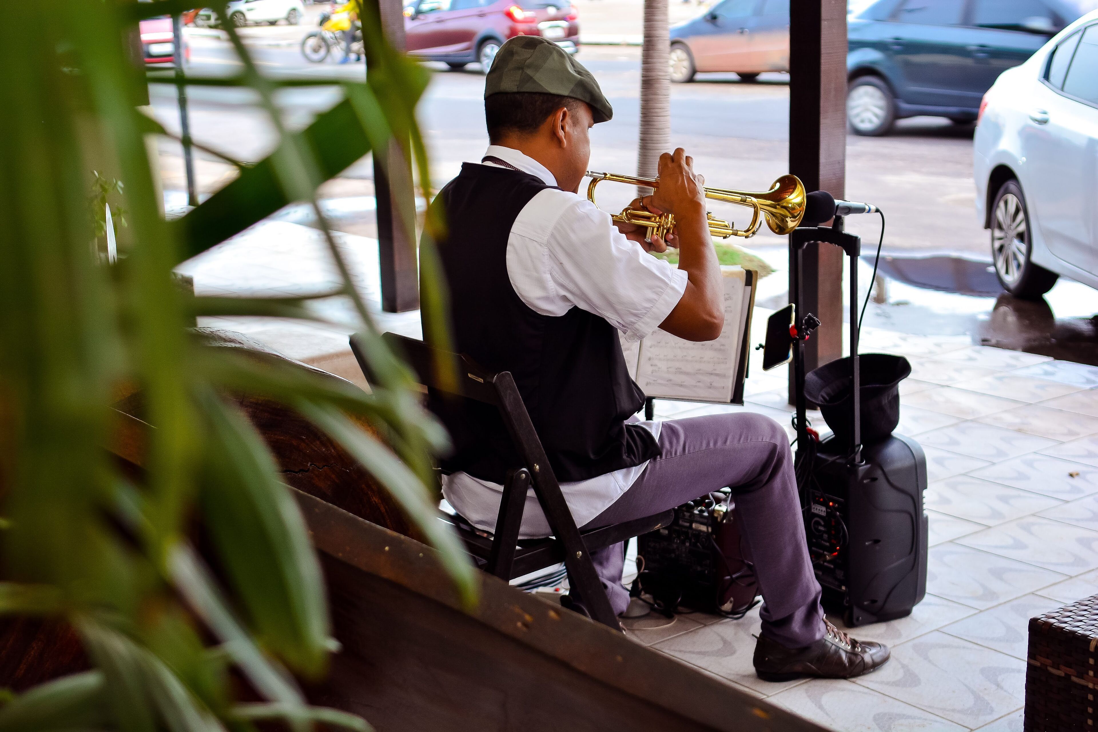 A person seated on a bench playing a trumpet, with a microphone and a suitcase nearby.