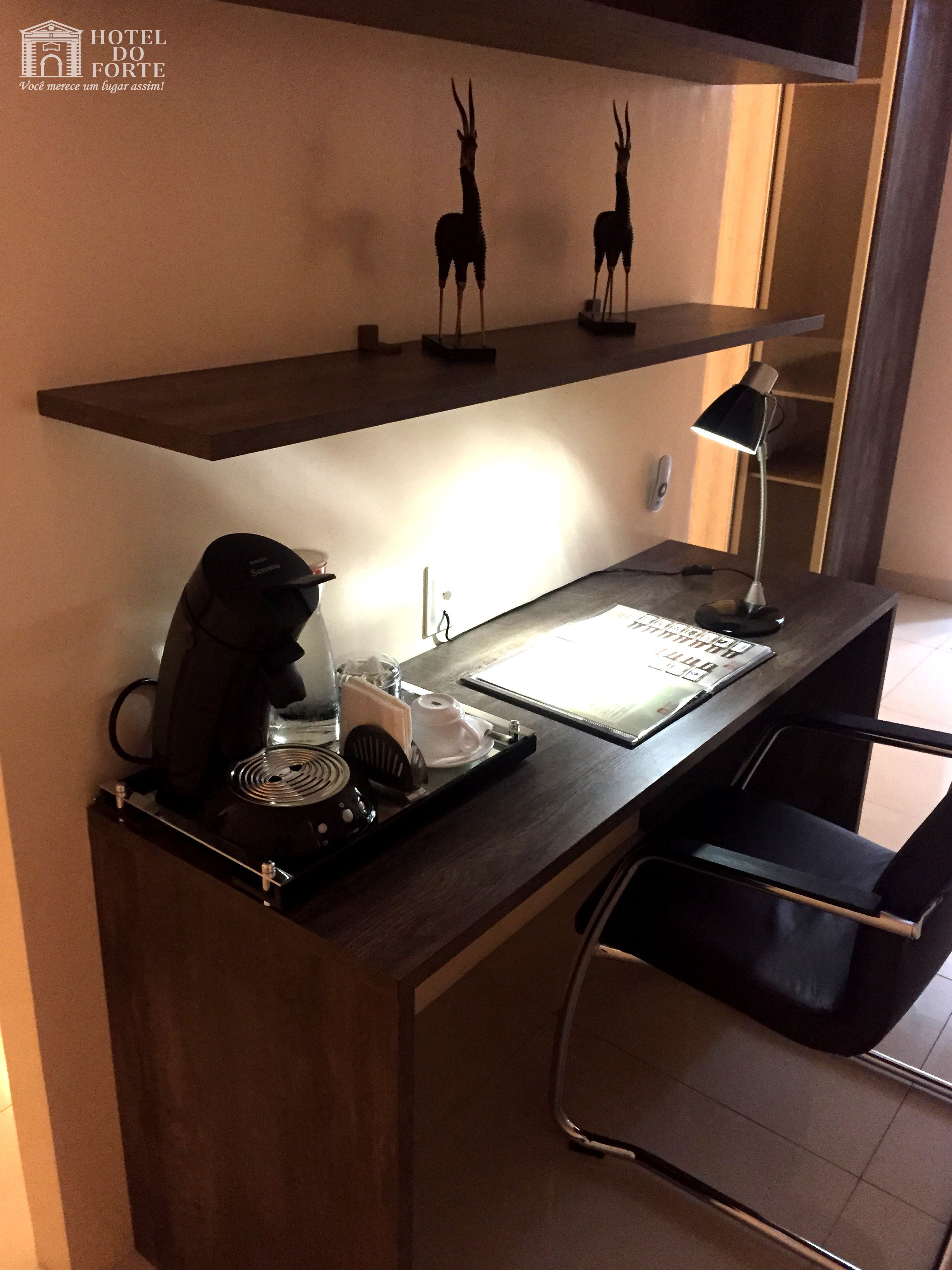 A hotel desk with a coffee maker, a lamp, and a tray with a notebook and pen. There are two decorative animal figures on a shelf above the desk.