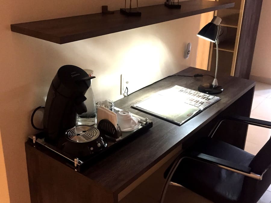 Escritorio de hotel con cafetera, lámpara y bandeja con cuaderno y bolígrafo. En una estantería sobre el escritorio, hay dos figuras decorativas de animales.