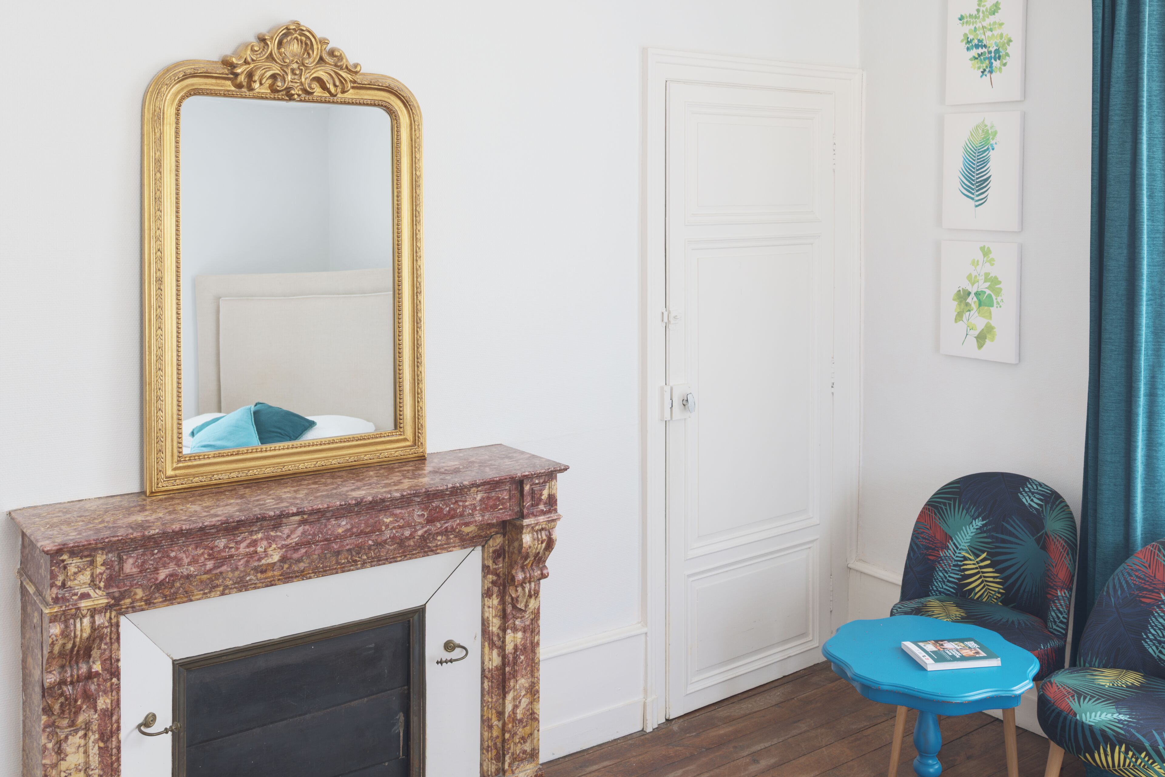 Une pièce avec une cheminée en marbre, un grand miroir décoratif, une porte blanche et un fauteuil bleu orné de motifs de feuilles tropicales.