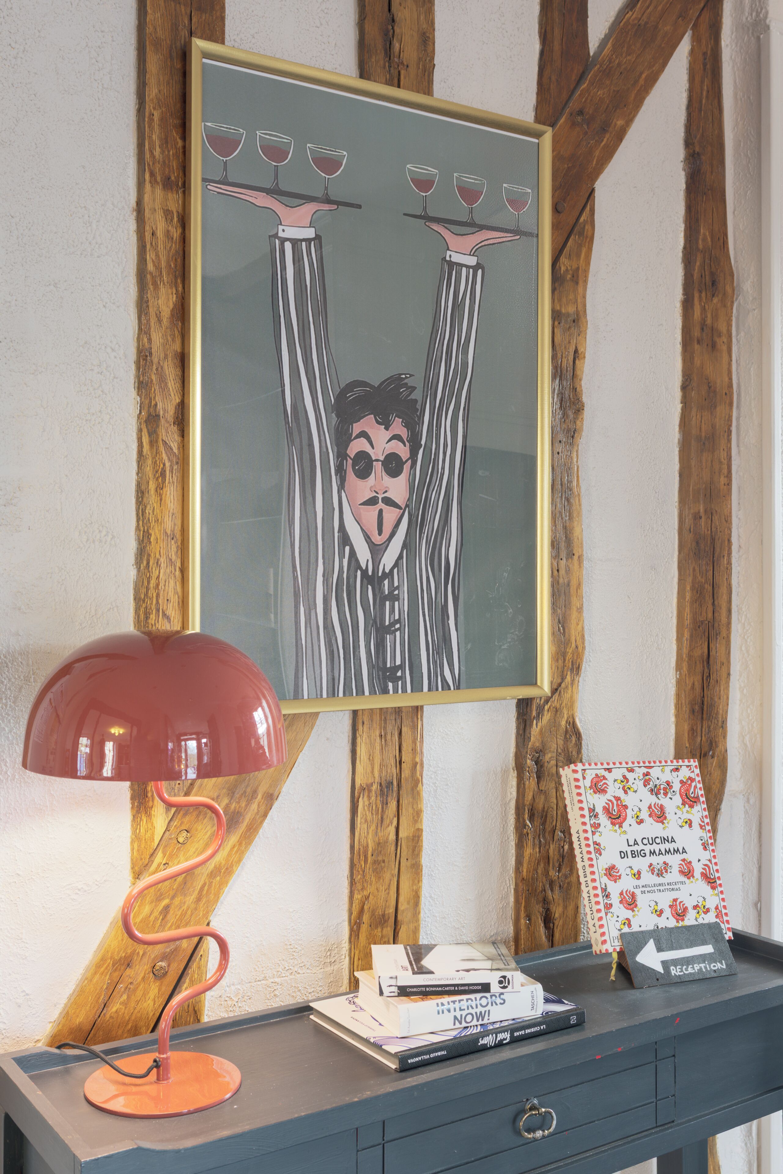 A framed artwork with glasses on a stand, a red lamp, a sign with an arrow, and some books on a table.