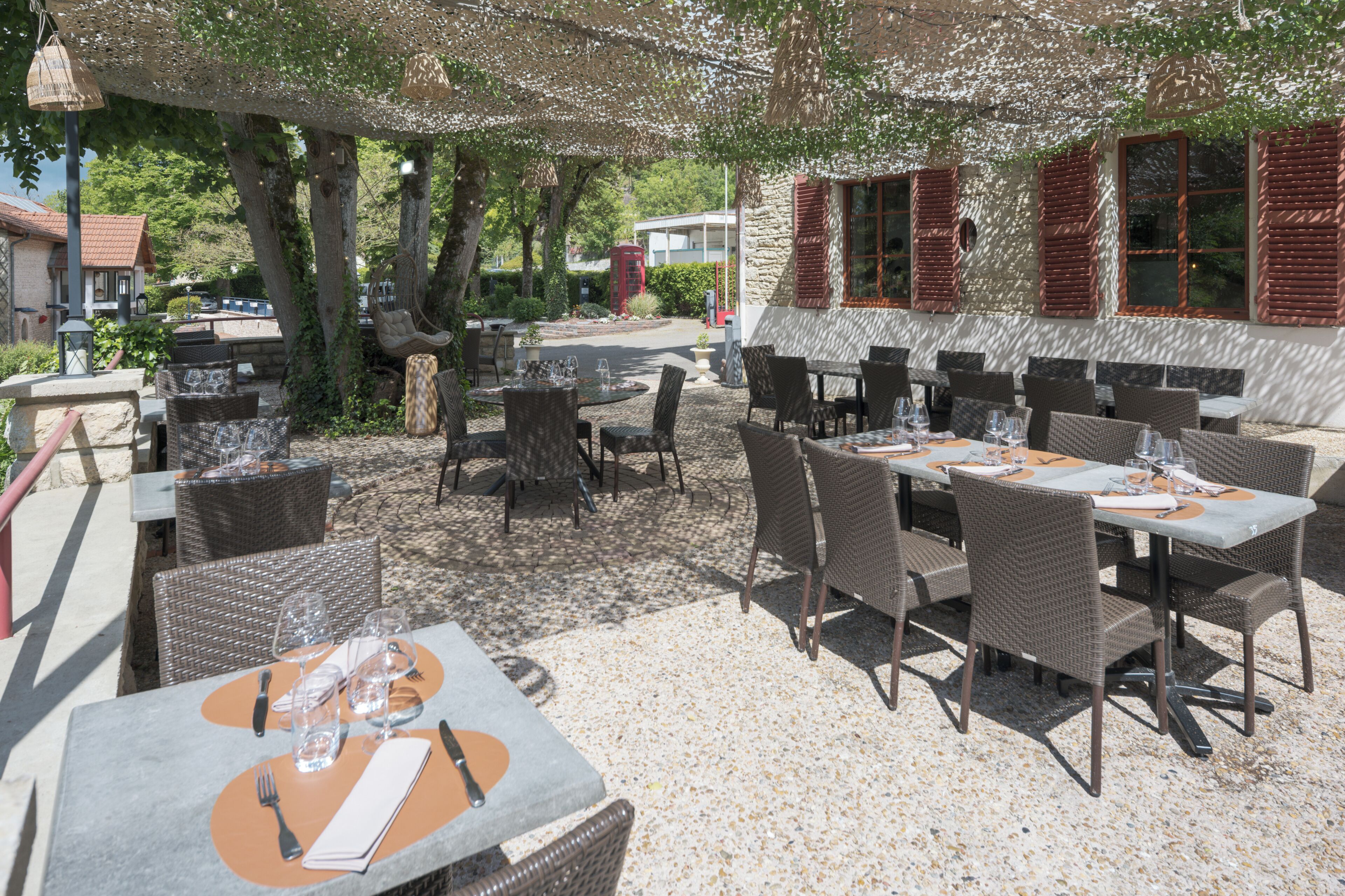 An outdoor dining area with tables and chairs, a stone building with red shutters, and trees providing shade.