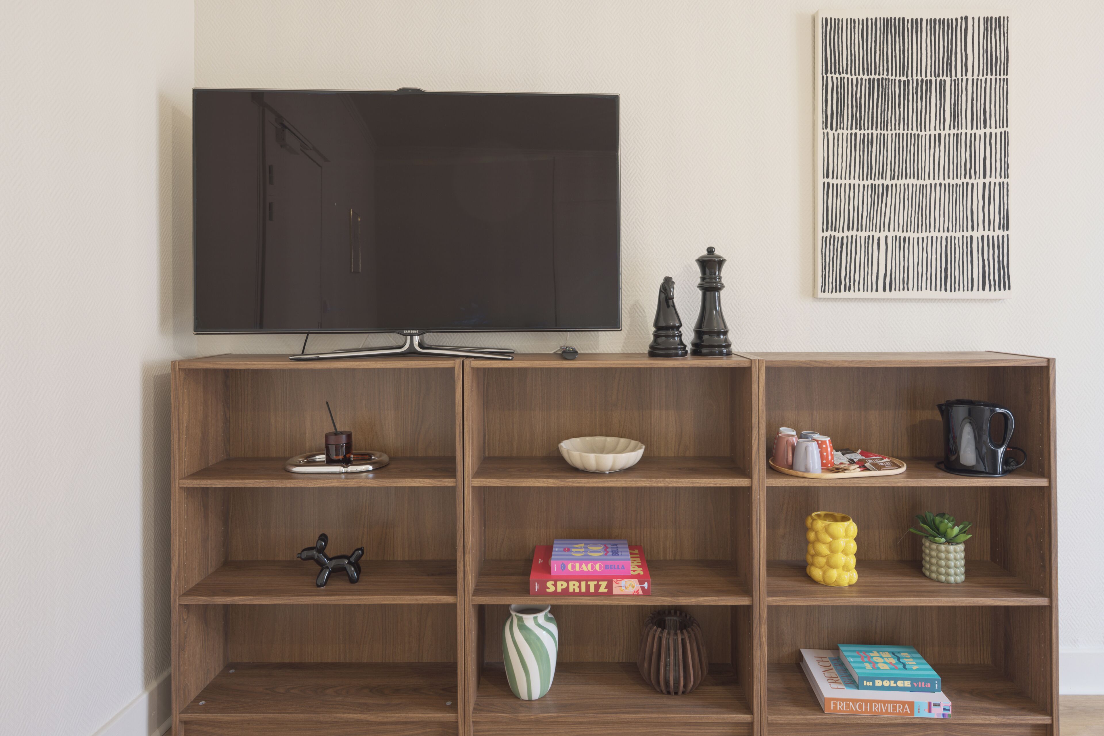 A wooden TV stand with a flat-screen TV, a chess set, books, decorative items, and a wall art piece.