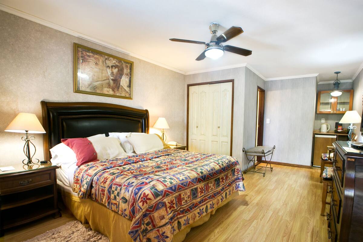 Ein Schlafzimmer mit einem großen Bett, zwei Nachttischlampen, einem Deckenventilator, einer Holzkommode, einem kleinen Tisch mit Stuhl und einer Tür zu einem anderen Raum.