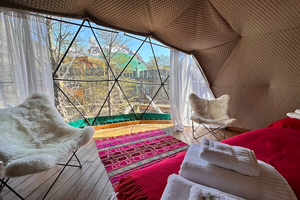 Una habitación acogedora con un ventanal grande, suelo de madera, alfombra colorida, una silla blanca y una cama con toallas blancas.
