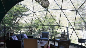 Estructura de domo geodésico con un sofá, sillas y una mesa en su interior, con vistas a un paisaje montañoso.