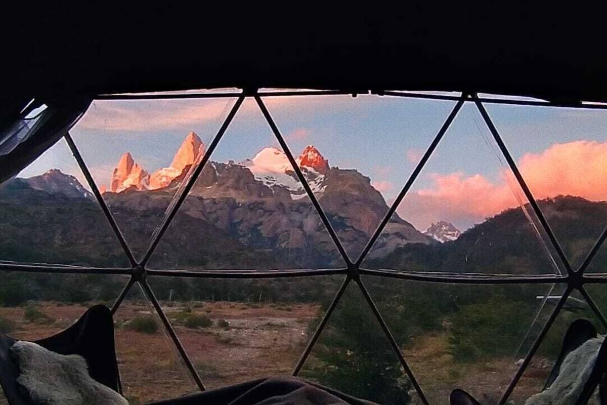 Vista a través de una ventana con estructura geométrica, mostrando un paisaje montañoso con picos nevados y cielo despejado.
