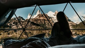 Una persona acostada en una cama con vista a montañas a través de una ventana con marco de metal.