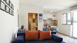 Uma sala de estar com um sofá azul, uma mesa pequena com toalha xadrez e uma cozinha ao fundo.