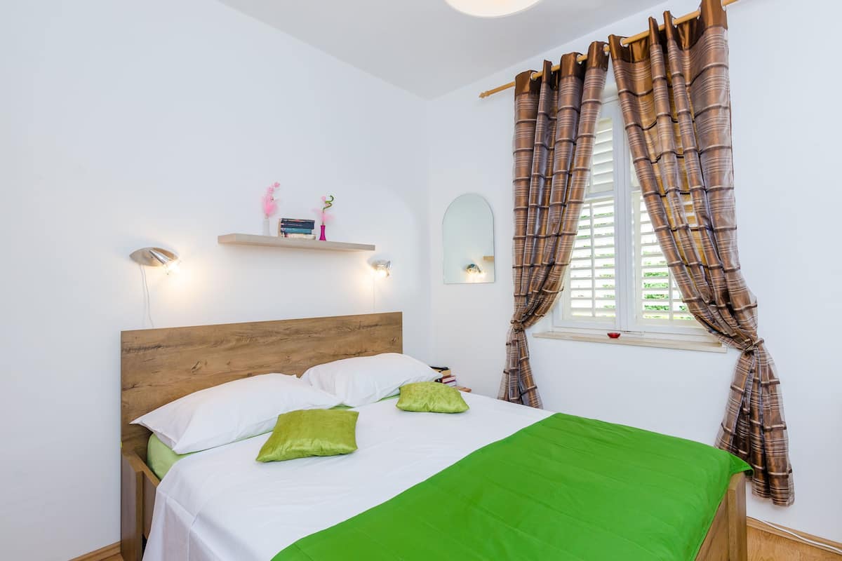 Cama bem arrumada com cabeceira de madeira, colcha verde e duas almofadas. Há uma estante com objetos decorativos acima da cama e uma janela com cortinas castanhas.