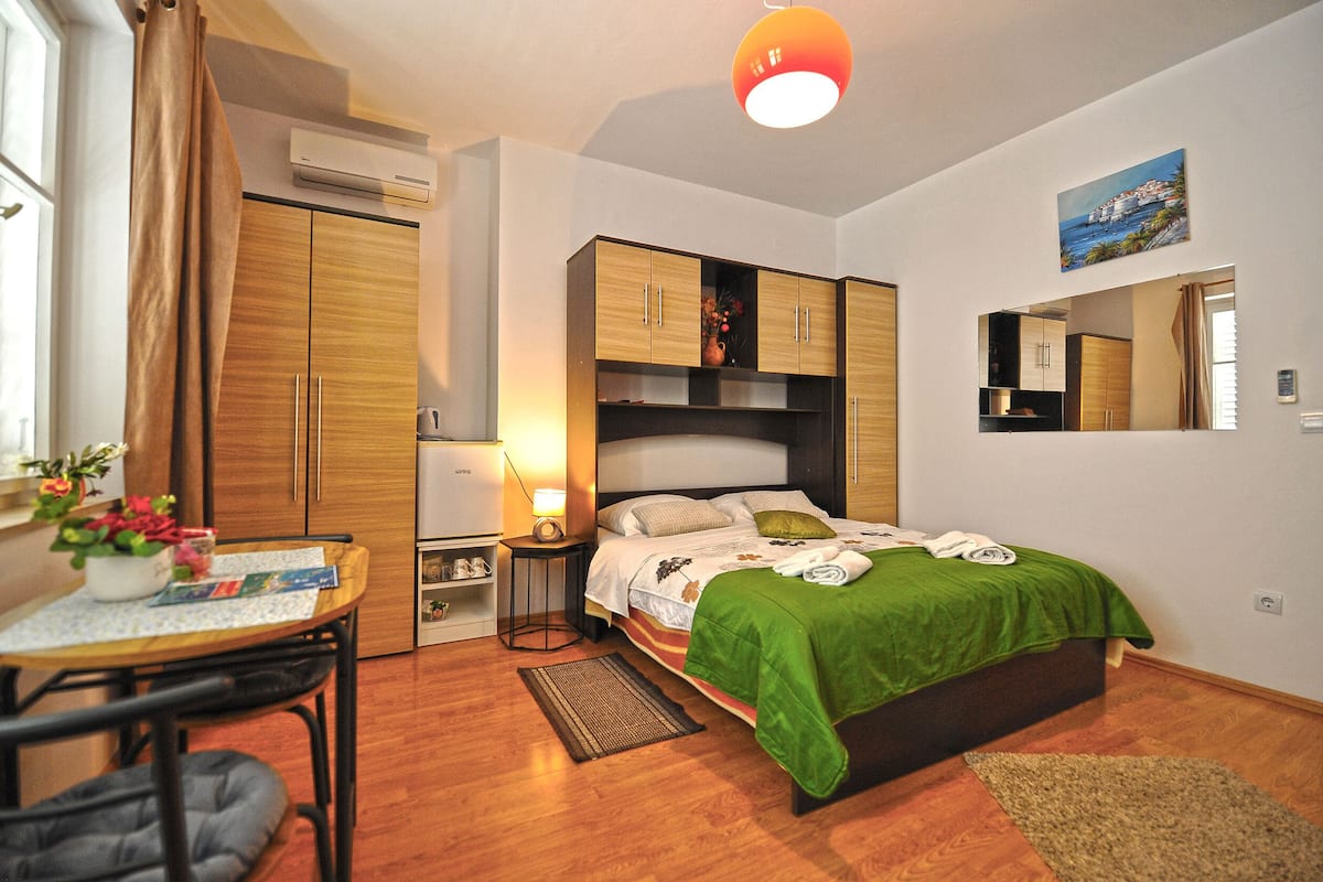 Quarto com cama, mesa, cadeira, escrivaninha, espelho e janela com cortinas.