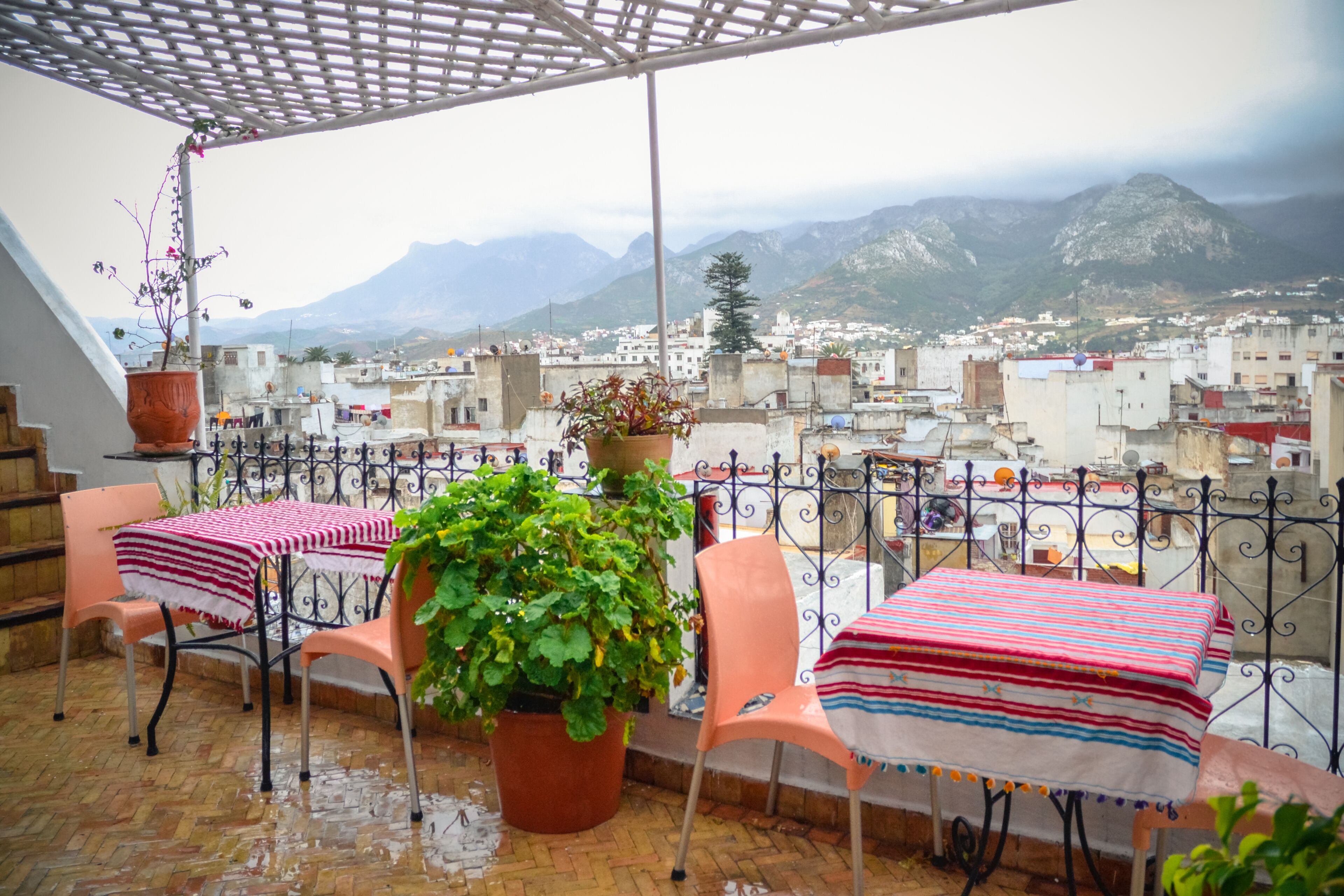 Una terraza con una mesa y sillas, una planta en maceta y vistas a un paisaje urbano y montañas al fondo.