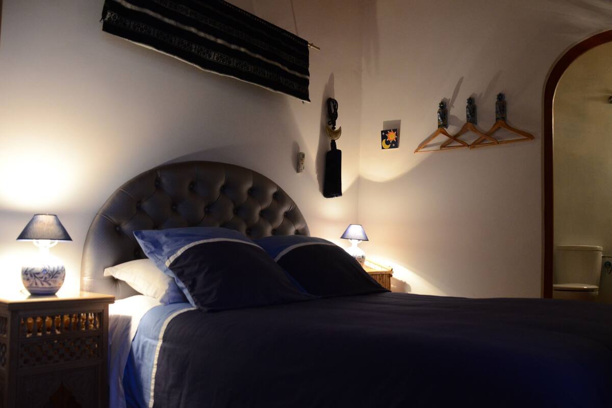 A bedroom with a tufted headboard, a bed with dark bedding, two bedside lamps, a wall-mounted air conditioner, and a mirror with a wooden frame.