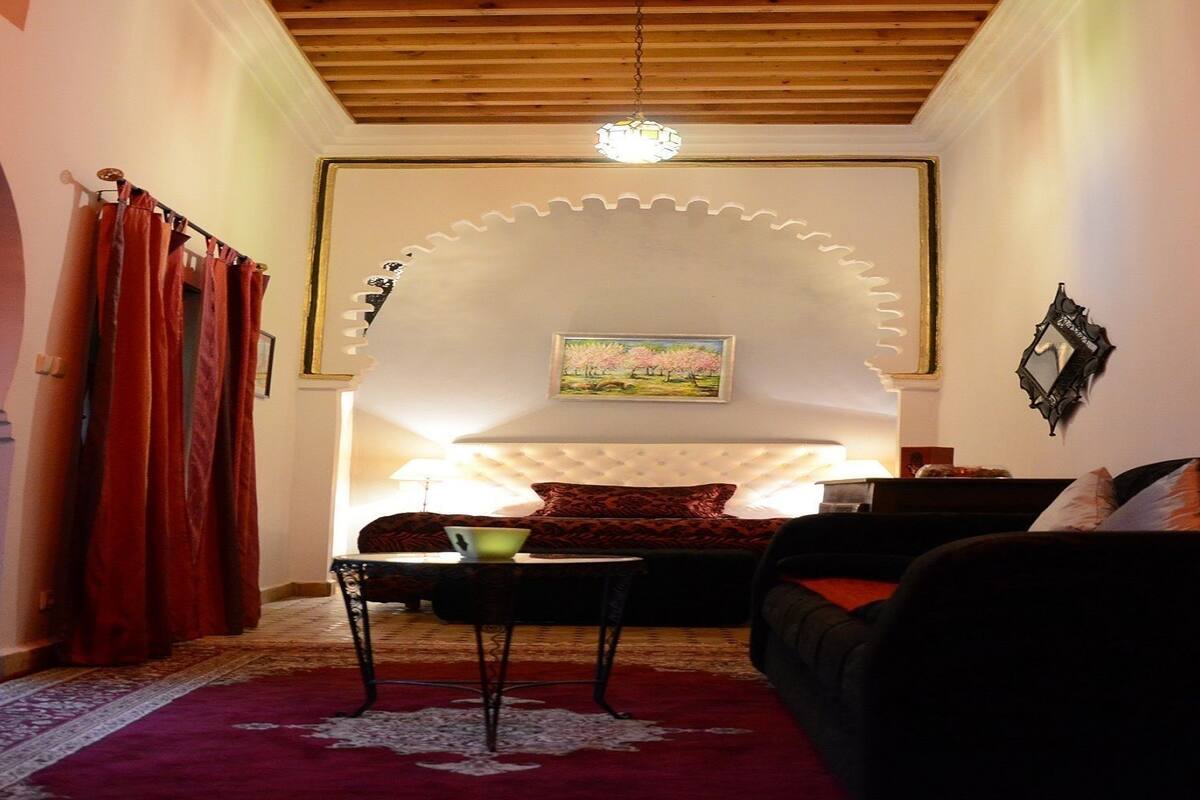 A room with a bed, a sofa, a small table, and a lamp. There is a painting on the wall and a window with curtains.