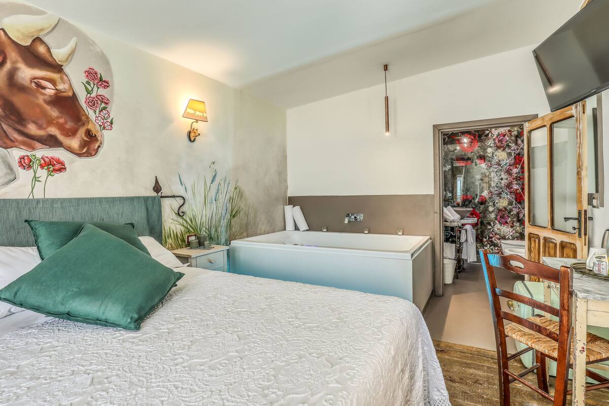 Une chambre comprenant un lit, un bureau, une chaise, un grand miroir, un téléviseur fixé au mur, et une salle de bain avec une baignoire et un lavabo.