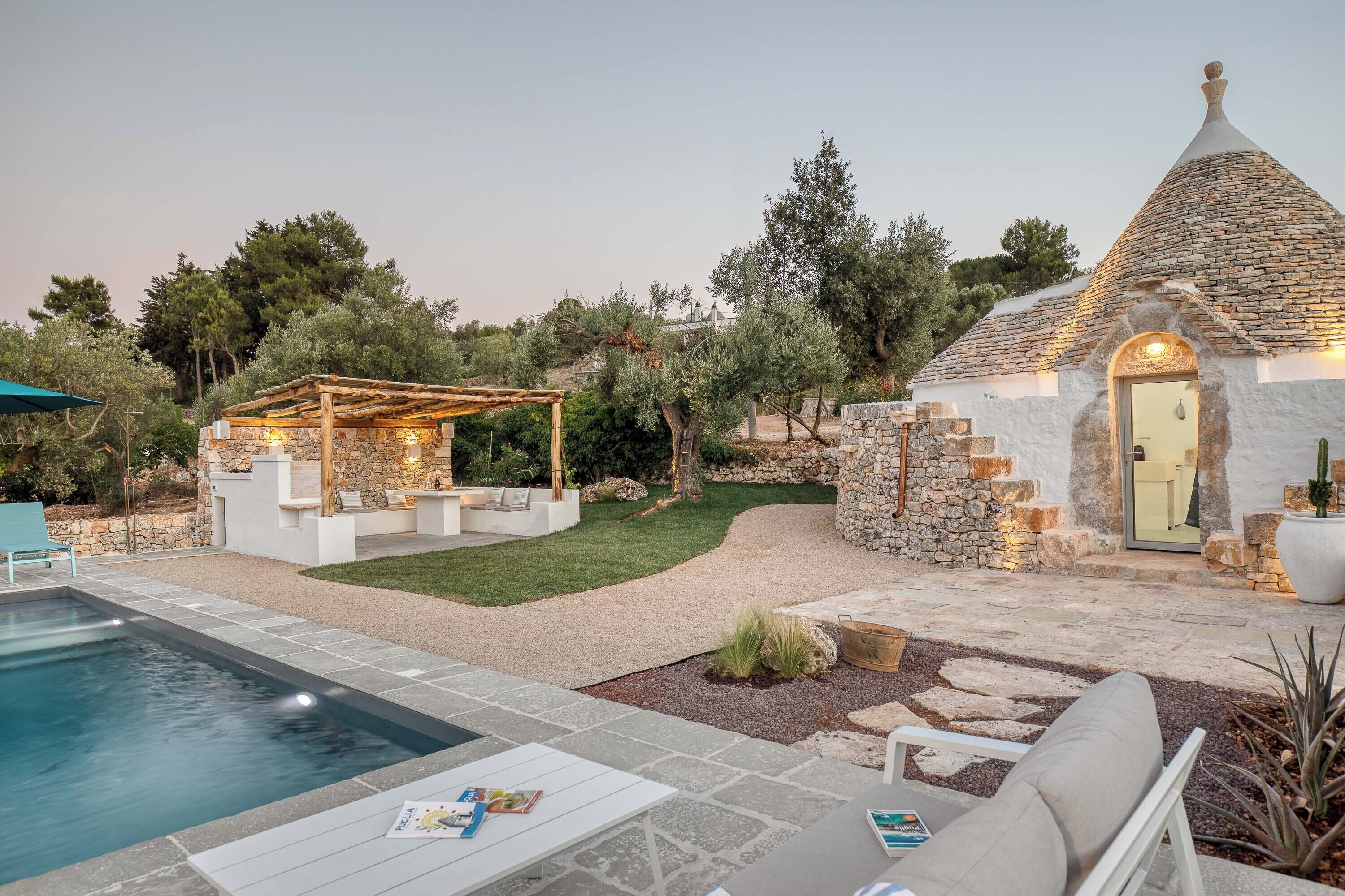 A pool area with a pergola, stone walls, and a small stone building with a door.